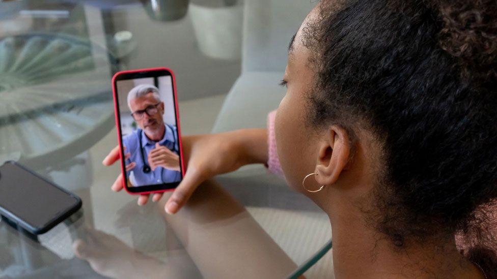 A woman, seen from behind, holds a phone and speaks to it. A doctor wearing a stethoscope round his neck is on the screen