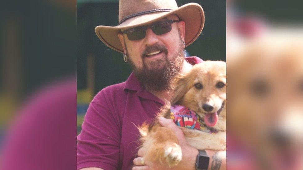 Matthew Young is smiling at the camera, wearing dark glasses, a beige cowboy hat and a purple polo shirt. He is holding a small rescue dog, called Leah. 