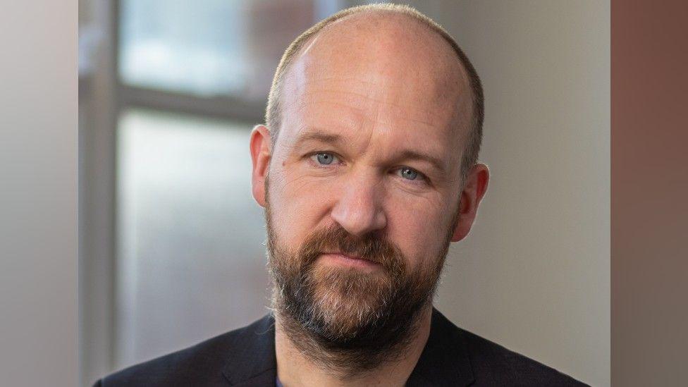 A headshot of Ian who has very short brown hair and long brown and grey facial hair. He is wearing a black jacket