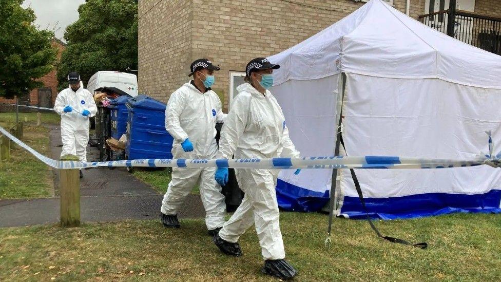 Three officers in white suits walking through a police cordon with a white tent.