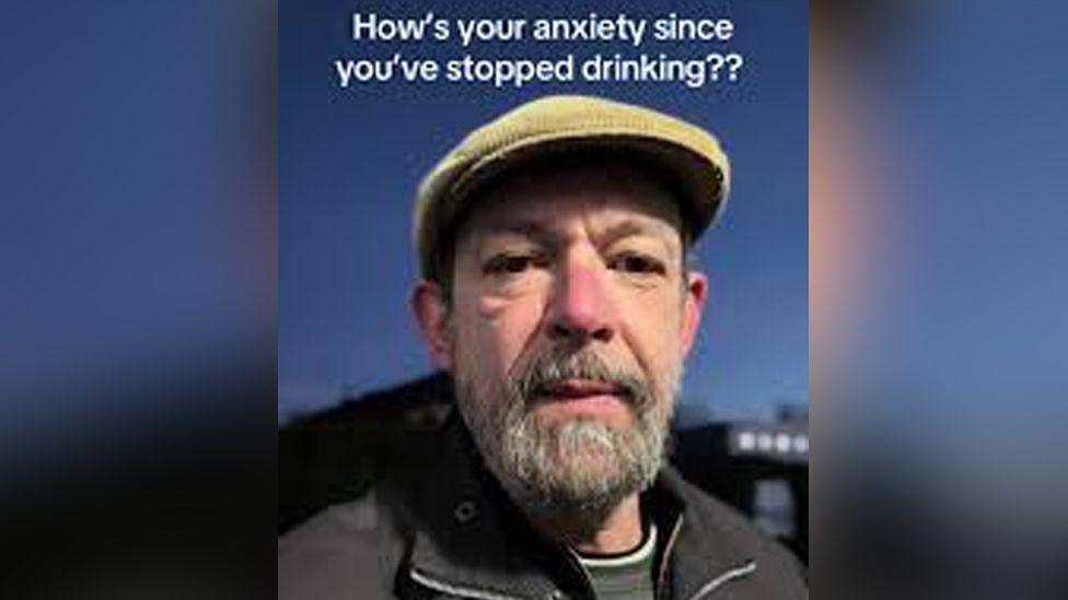 A still image from one of Mr Bone's TikTok videos. He is wearing a coat and a hat. Text in the upper quarter of the image poses the question "how's your anxiety since you stopped drinking??"