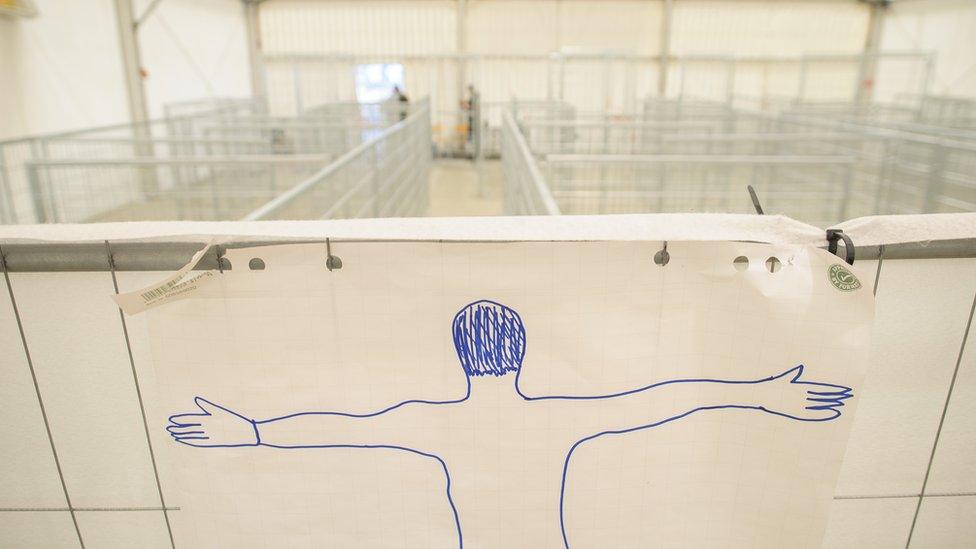 A crudely drawn marker image shows a person spreading their limbs, apparently ready to be searched - behind it is a large tented area of metal barriers - but no people