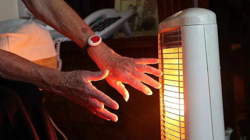 Close up shot of someone warming their hands in front of a glowing electric heater. A telephone and a family photograph can be seen in the background.