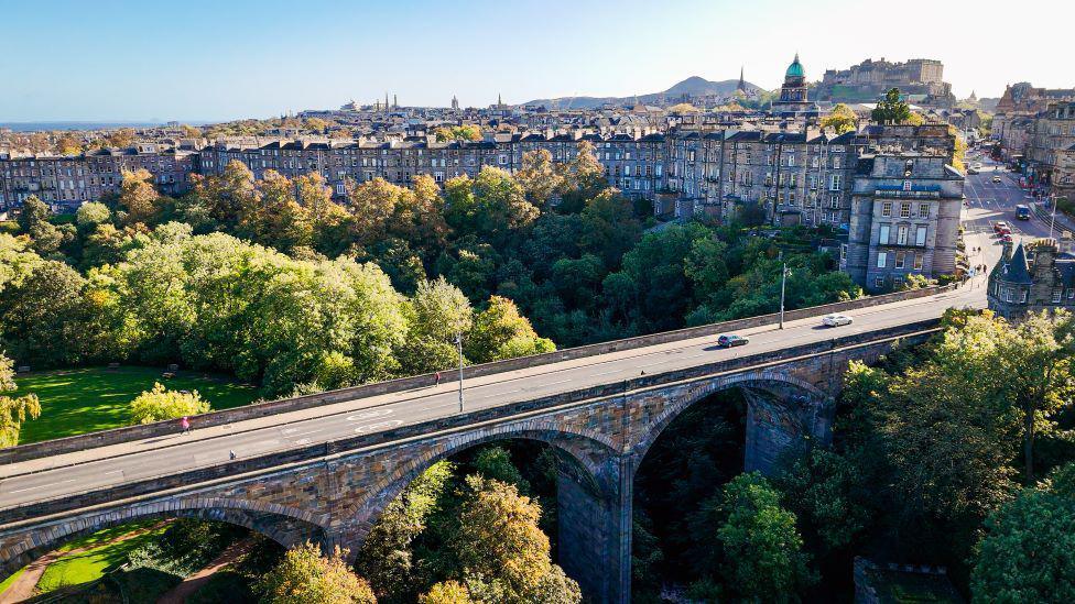 The Dean Bridge made up of large arches with a road over the top, It is leading to a number of old flats and underneath there are lots of green trees.