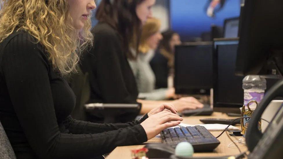 A close-up of a woman with curly blonde hair sitting in a row of people who are at a desk typing on keyboards.