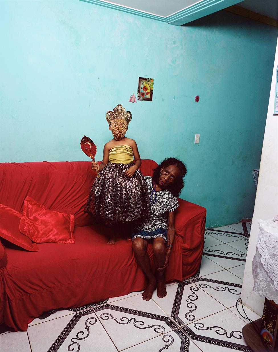 A photo by Deana Lawson of a child wearing a headdress stood on a red sofa, next to a woman who is sitting down