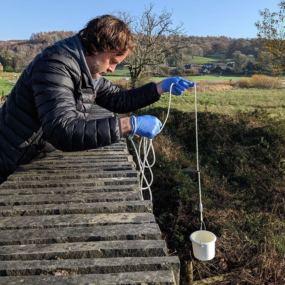 Ged Dolan leaning over a stone bridge with a bucket on a rope to collect water sample from the beck below