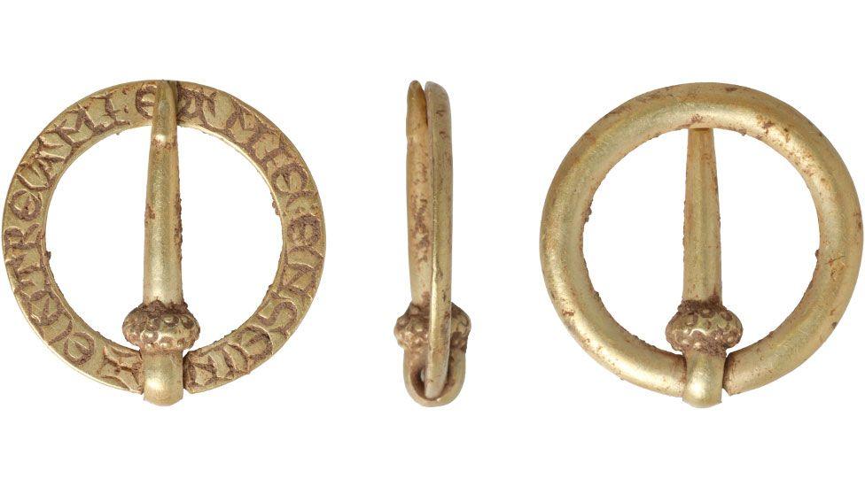 Three views of 13th or 14th Century brooch, made of a circular band of gold, showing the inscribed back, a side view and the plain curved front, with a pin, attached on one side of the band with a loop and swung across the middle to touch the opposite side of the band.