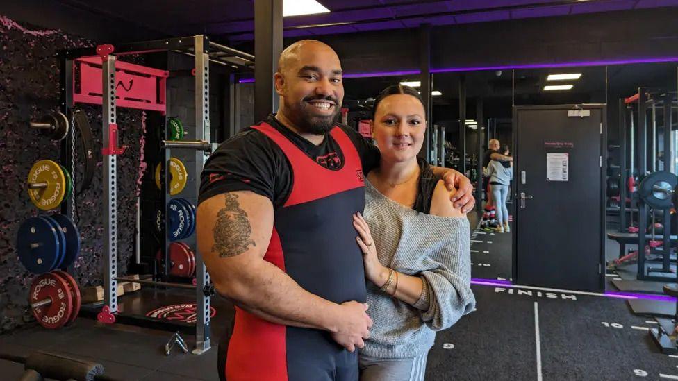 Ram Patten, in a gym, with an arm around his wife. There are weights and a squat rack behind him. He is wearing a red and blue top while she is wearing a grey jumper and has her hand on his chest