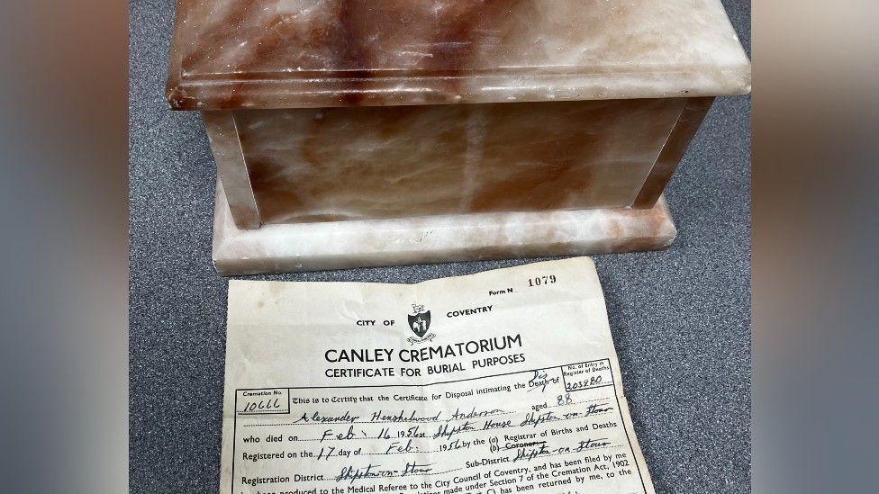 A brown and white marble box and yellowed burial certificate on a grey table top. The name Alexander Anderson and age of 88 can be made out on the certificate.