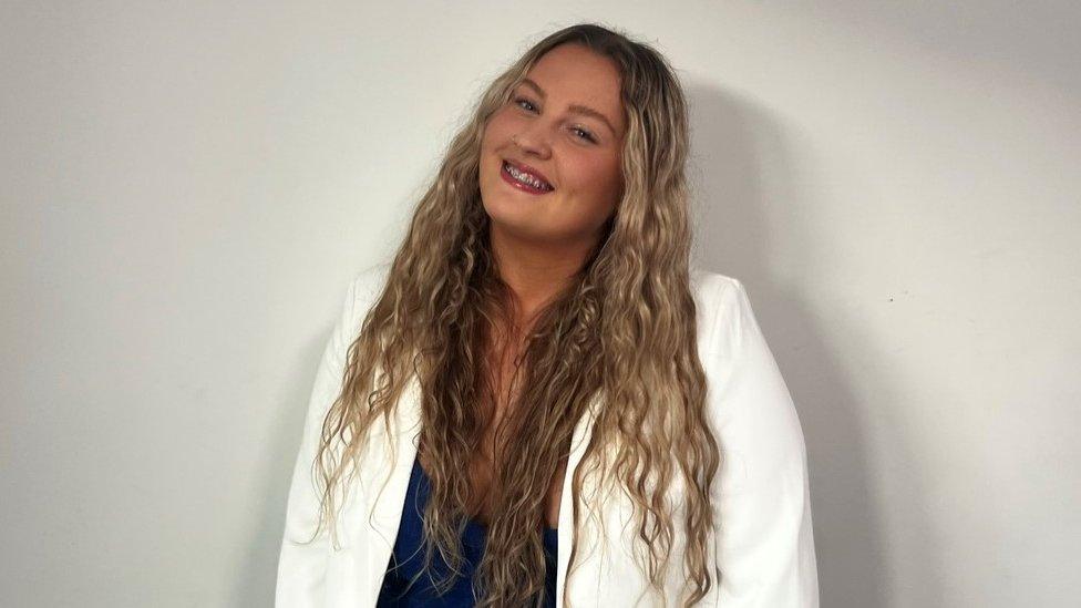 Mia Hardy smiles at the camera. She has blonde, wavy hair, and is wearing a navy blue dress with a white blazer.