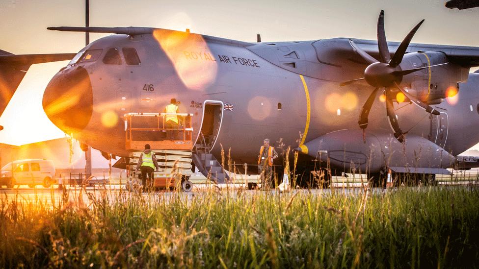 The Atlas A400M on the runway, with staff bustling around it, and a side hatch open, with steps leading down. The sun is behind the plane, creating a golden lens flare.