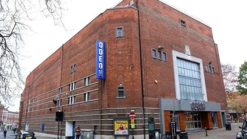 The cinema is a giant red bricked building. The silver Odeon logo is above the entrance. A vertical sign is fixed higher up on the edge of the building.