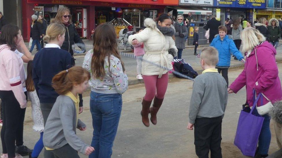 Colour photo of people skipping in a street near shops