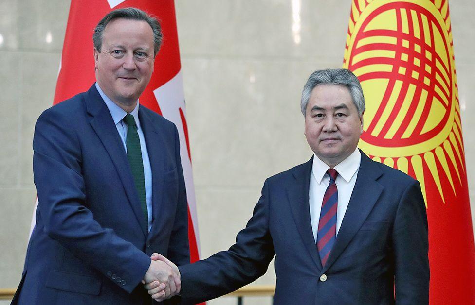 Mr Cameron (L) and Mr Kulubaev (R) shake hands while standing in front of the British and Kyrgyzstan flags. 