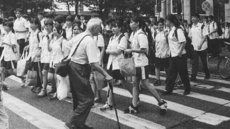 An older man walks across a zebra crossing with lots of school girls walking the other way