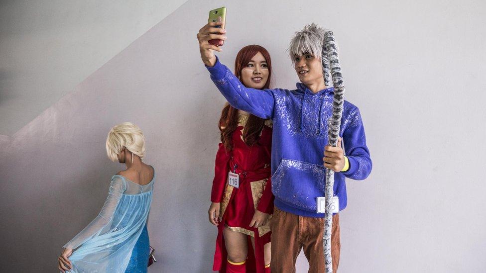 Male "cosplayer" in blue takes a selfie of himself and a woman in a red dress, at the event