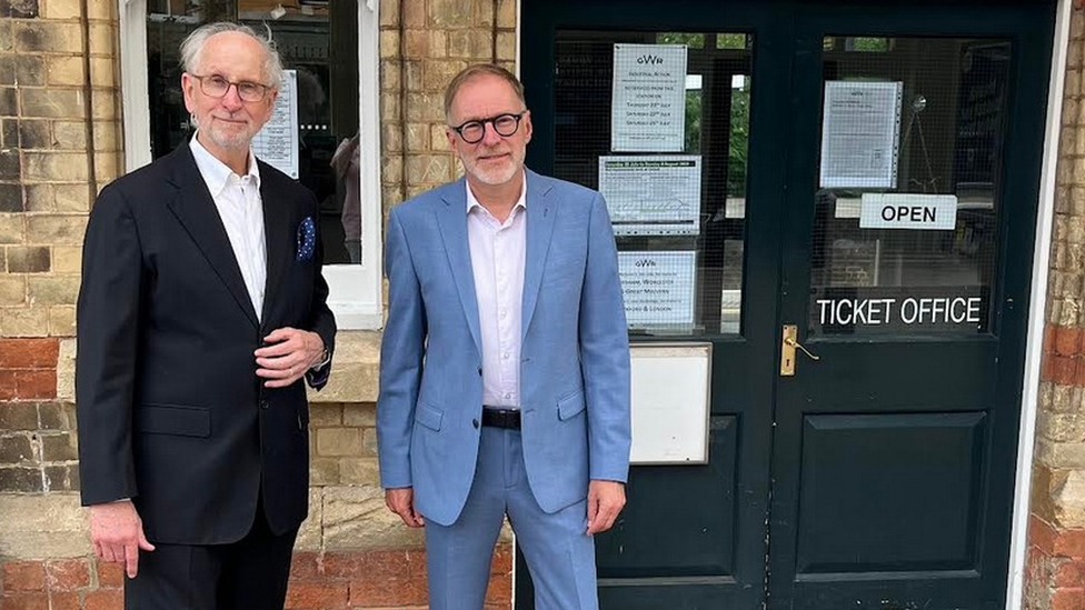 Two men wearing suits standing next to a ticket office