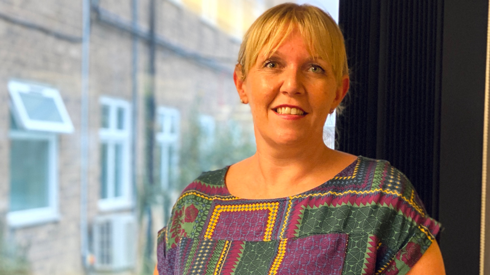 Deborah Needham with short blond hair smiling at the camera and wearing a green, purple and yellow dress. There is a window behind her looking out onto a brick building with white window frames