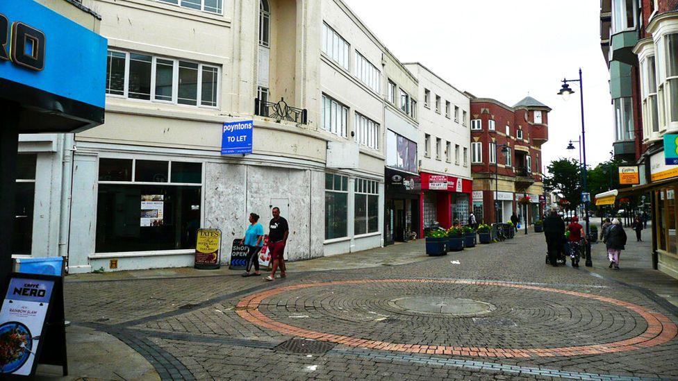 People walking along Strait Bargate, a narrow, pedestrianised street, in Boston. Shops and cafes including Cafe Nero and Cooplands can be seen. Other shopfronts are empty