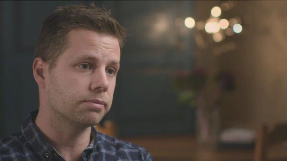 Nick Bailey, wearing a blue shirt, looks off camera in a dark room