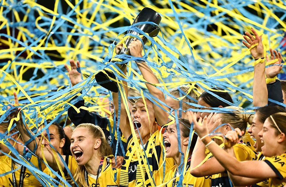 Taren King of Central Coast Mariners lifts the A-League Women's Grand Final trophy after their penalty shootout win over Melbourne Victory, with players covered in colourful tickertape. Photo by Quinn Rooney