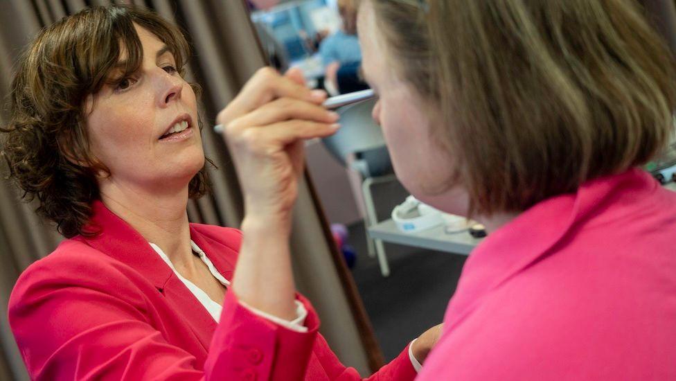 A woman applies make-up to her younger colleague. Both women are both dressed in pink and have shoulder-length dark hair.