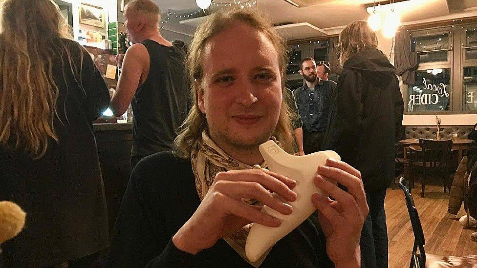 Robert Hickman sitting at the Greenbank Pub. He has shoulder length hair and is holding is home-made ocarina in his hands.