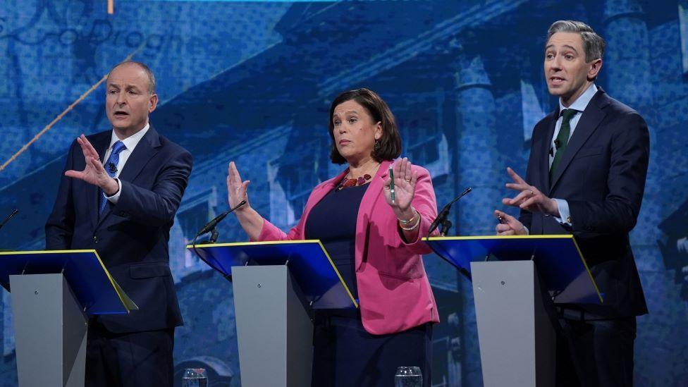 Micheál Martin stands to the left in front of a grey podium. He is wearing a blue suit and tie and is holding his hand out. Mary Lou McDonald stands in the middle in a blue top and bright pink blazer, holding two hands up. Simon Harris stands behind a podium to the right and also appears to be in the midst of talking. 