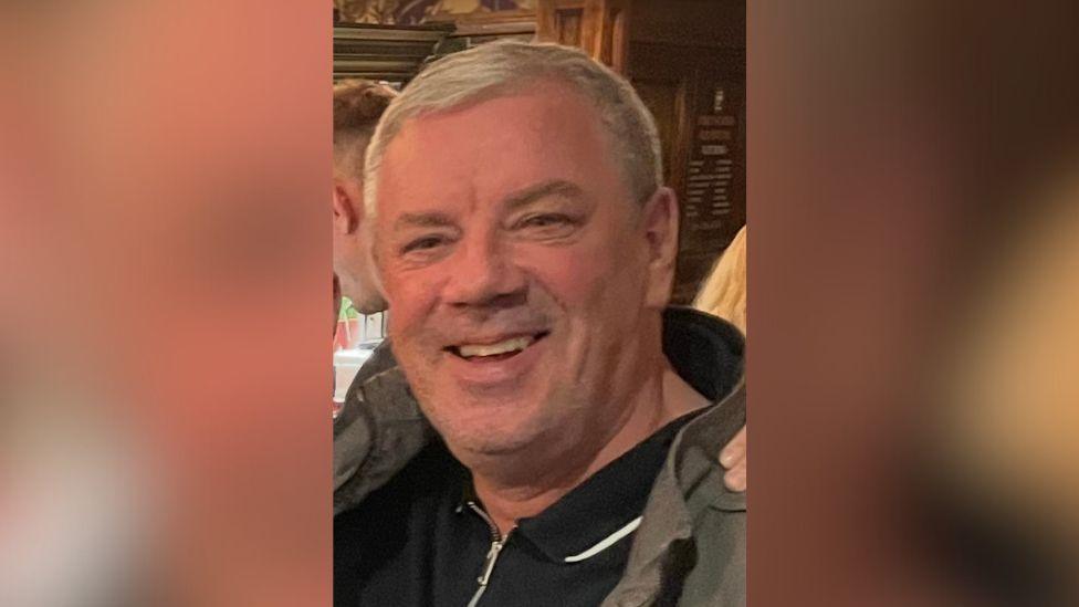 A man with grey hair, wearing a green jacket and black polo shirt underneath is stood at what seems to be a bar or restaurant. He is smiling and looking directly at the camera