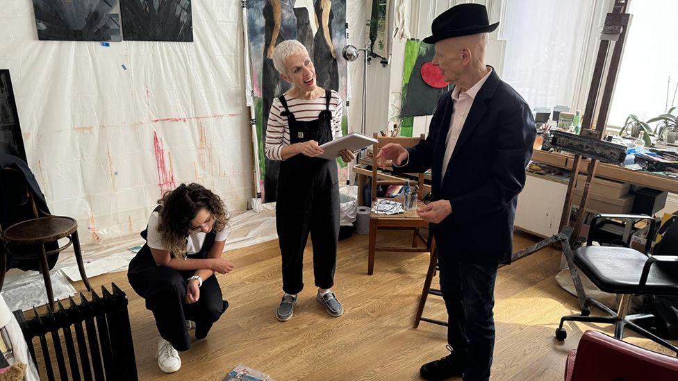 David Remfry in his studio wearing a suit and black hat talking to curator Hannah Willetts, who has white hair and is wearing black dungarees. Another woman is crouched down looking at his work and there are paintings on the wall