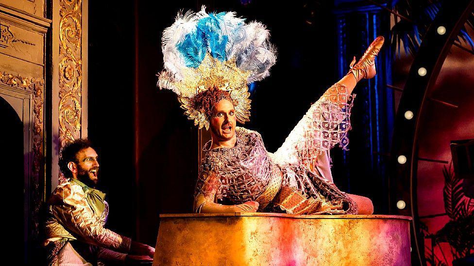 Seiriol Davies is on top of a grand piano in a brown and gold dress, which gives the impression of a Roman soldier or Egyptian ruler. They wear an elaborate headdress of gold segments topped with peacock blue and white feathers. Another performer wearing a gold jacket sits playing the piano. Both appear to be singing.