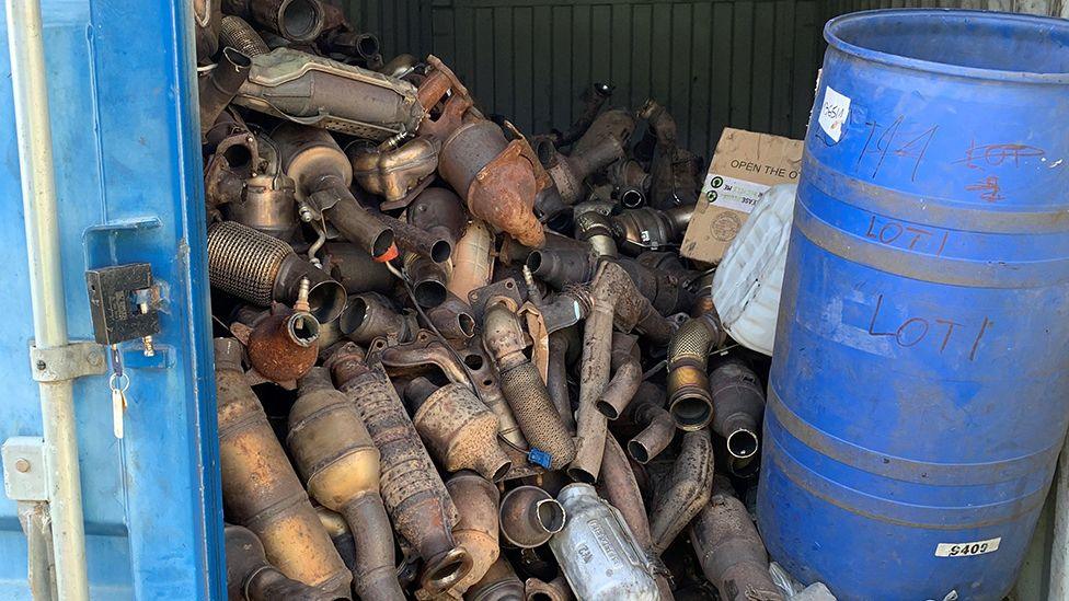 A blue shipping container filled with rusting catalytic converters and a blue plastic barrel