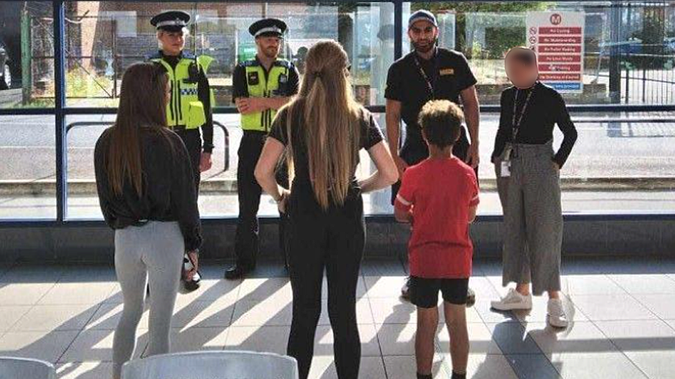 Rashid Zaman is wearing a black cap and black T-shirt. He's shown alongside two uniformed community police officers, and a woman in grey trousers, as they converse with three children children at Ossett bus station in West Yorkshire