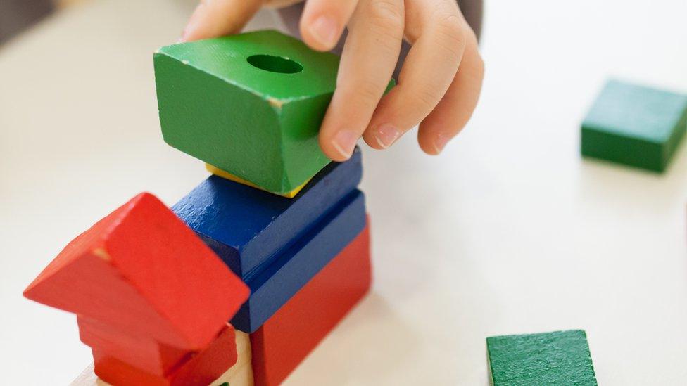 Child's hand playing with coloured wooden brick shapes on white table