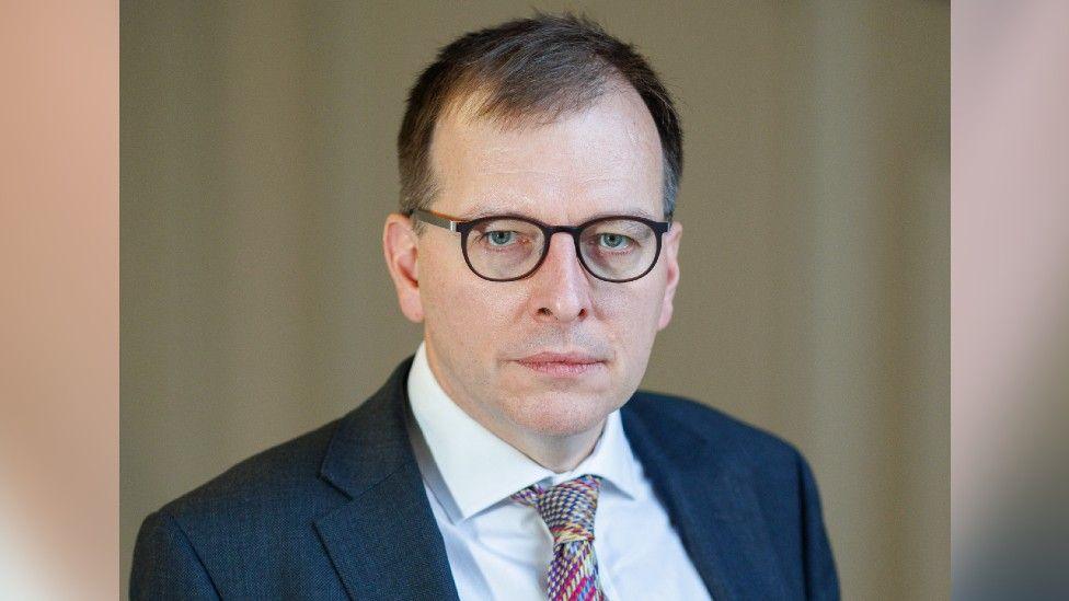 Tom Roques a man who is wearing glasses and looking directly at the camera. He is wearing a navy suit jacket, white shirt and a patterned coloured tie.