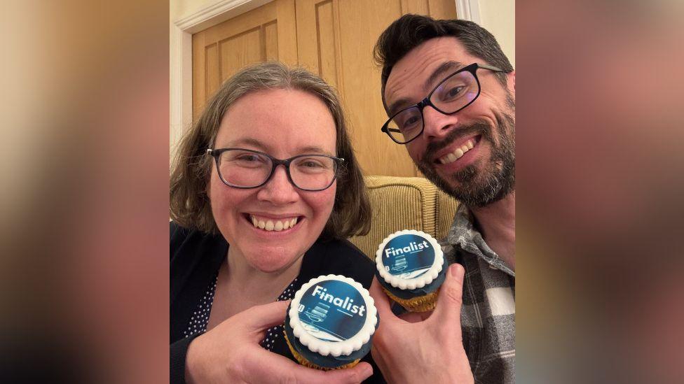The two people from the above photograph, this time sitting on a sofa, grinning at the camera and each holding up an object reading "Finalist".
