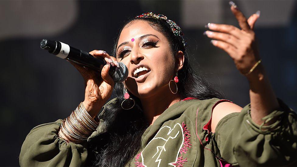 Raja Kumari, a female artist, wearing a green outfit, singing into a black microphone with her left hand pointing outwards.