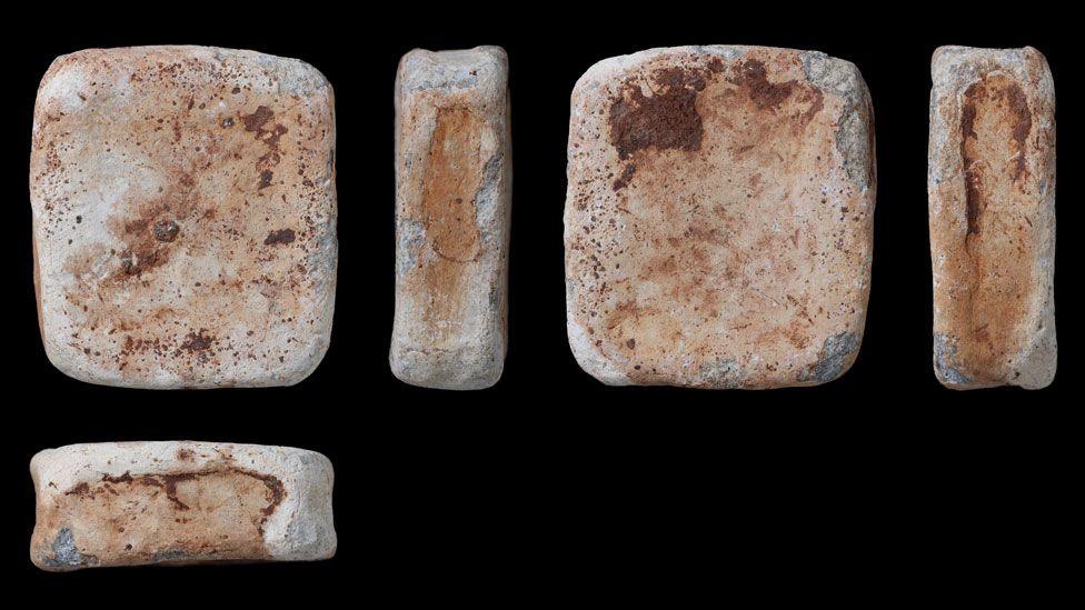Five views of a lead ingot. It is creamish in colour and speckled with brown. Four views along the top of the picture show the rectangular block with rounded corners; the second view is on its side; the third view is its underside; the fourth view is one of its sides. The fifth view is under the first and shows a third side.