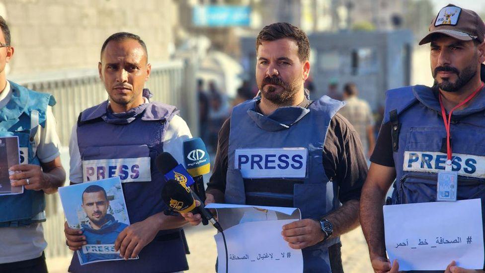 Four Palestinian journalists are pictured, all of them wearing blue flak jackets with the word "press" on them. They are holding pictures of Hamza Murteca in his memory. It appears to be a sunny day and they are all standing on a street in Gaza.