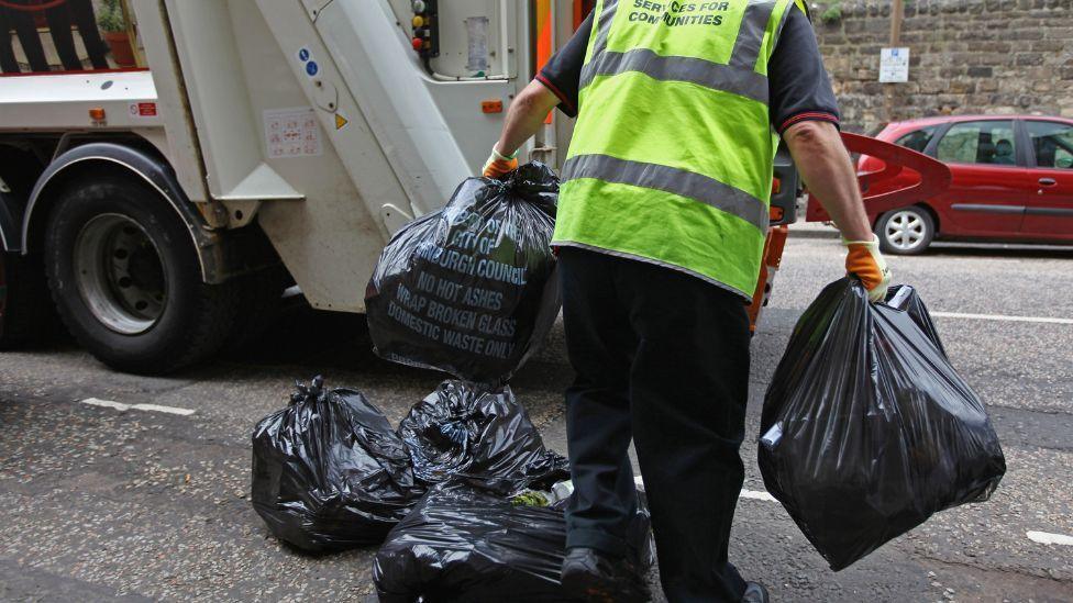 A man in a high vis jacket carries black bin bags to a bin lorry 