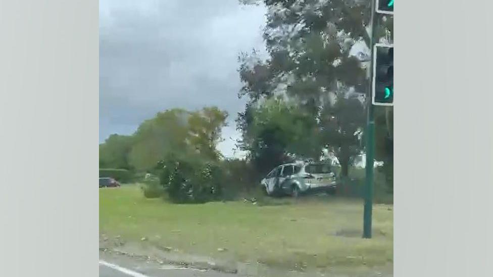The white car on the roundabout. It has hit a tree, which is damaged. The car is also badly damaged. It is covered in black scorch marks and its windows are smashed.