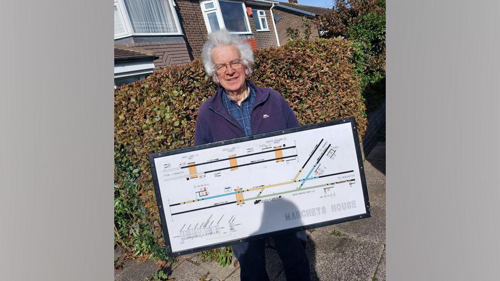 A member of the Northumberland Line Heritage Community Group holding a diagram from Marcheys House. The diagram shows lines representing the track. They changre from black to green, blue and yellow. The man holding he diagram is wearing a fleece and has grey hair.