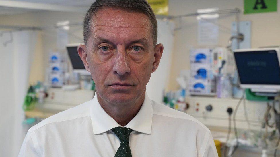 Matthew Hopkins wearing a black tie and white shirt inside a hospital ward. He has short brown hair and looks serious in his expression.