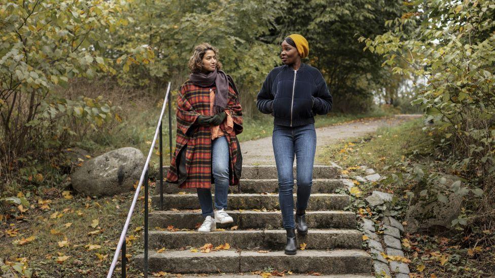 Two women, one in a long red plaid coat, jeans and a scarf, the other in a black coat, jeans and a yellow beanie, walk down stairs in a park and talk, surrounded by autumnal leaves