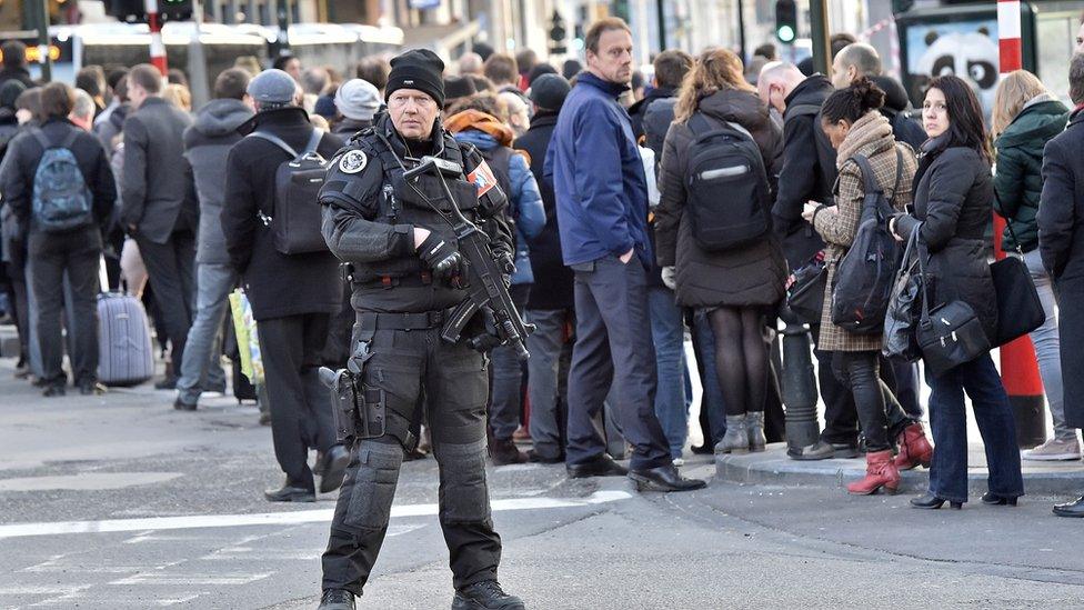 An armed police officer oversees queues of people waiting for a bus