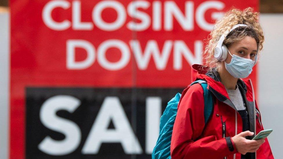 Woman wearing face mask and headphones walks past a closing down sign in a Cardiff shop window