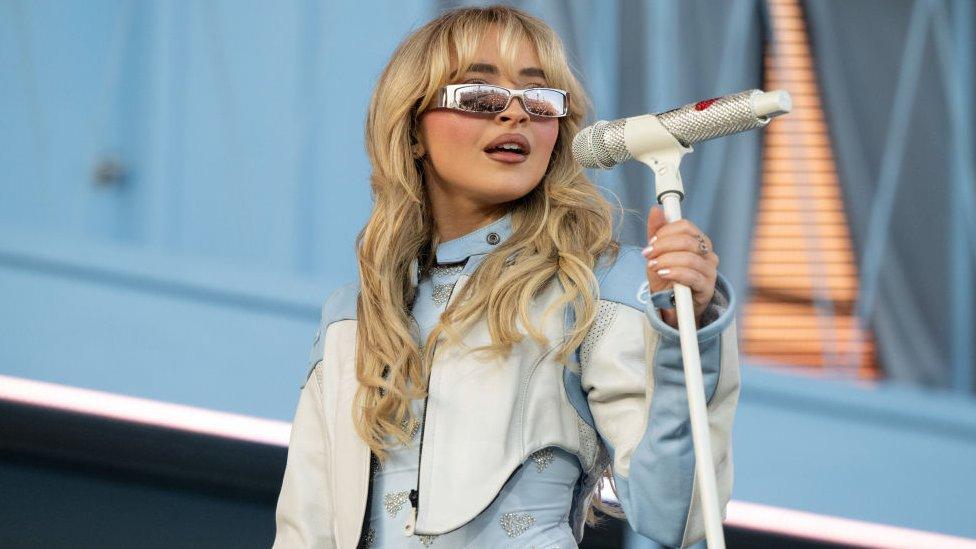 Sabrina Carpenter on stage at Coachella, she is wearing sunglasses and a blue dress and jacket