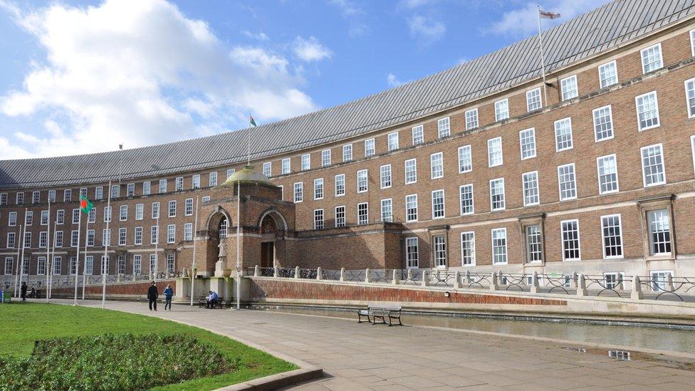 City Hall, Bristol. It is a wide, brown brick building with lots of windows and flags. A green area and people can be seen in front of the building.