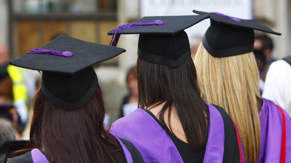 A group of three femal graduates, two with dark hair and one with blond hair, pictured from behind wearing robes and square academic caps